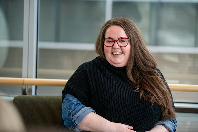 Kelley Jones sitting in the HUB at Mass General Cancer Center, wearing red glasses, smiling.