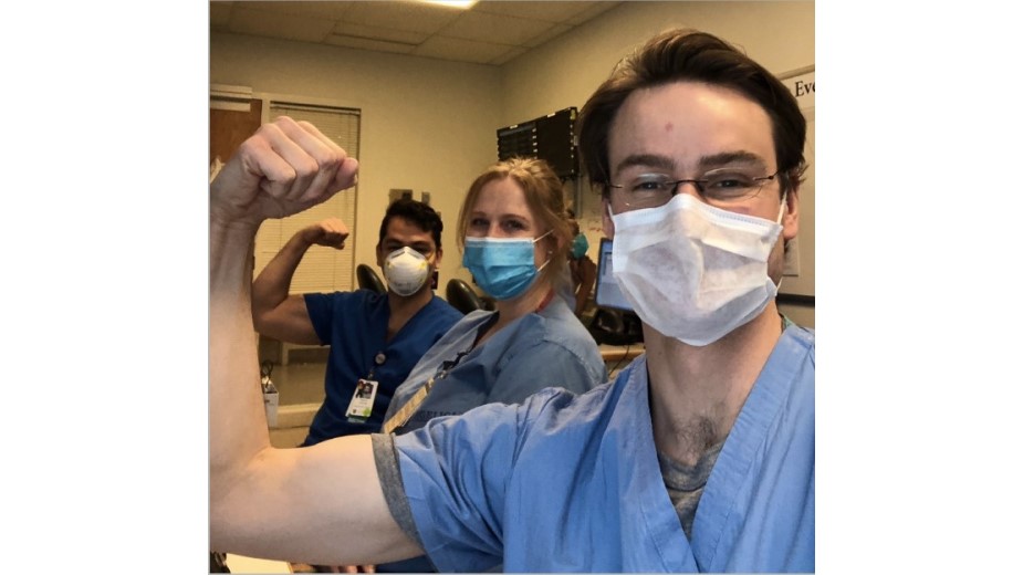A young man in mask and scrubs flexes his bicep. His arm frames another young man and woman in mask and scrubs. A young man in mask and scrubs flexes his bicep. His arm frames another young man and woman in mask and scrubs.